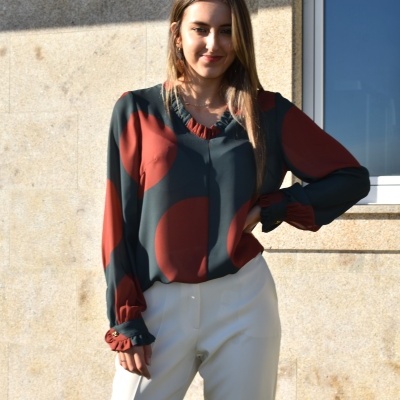 Blusa feminina verde escura com manchas vermelhas, gola em V com franzidos e punhos elásticos, usada com calças brancas, ao ar livre junto a parede de pedra e janela.