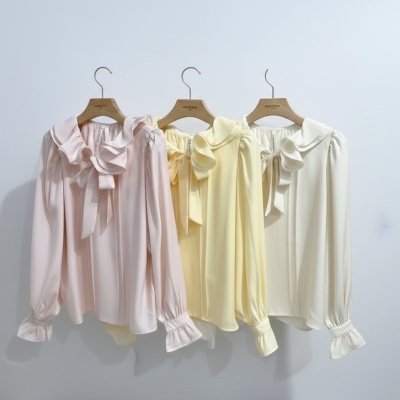 Três blusas femininas em cores pastel com babados penduradas em cabides de madeira