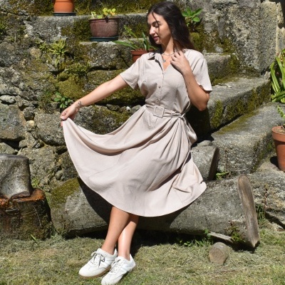 Mulher vestindo vestido bege de tecido leve com cinto e ténis brancos sentada em degrau de pedra ao ar livre