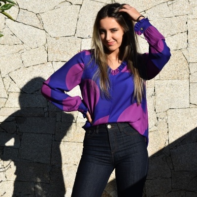 Blusa feminina azul e roxa com calças pretas em fundo de parede de pedra