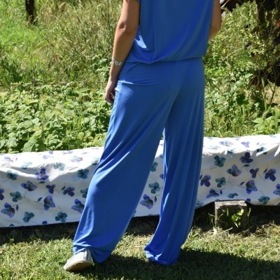 Conjunto de roupa azul fluído com camiseta e calças largas, pessoa de costas em relvado diante de vegetação e tecido floral.