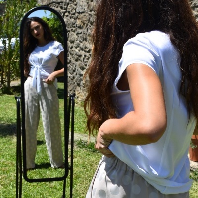 Mulher com t-shirt branca e calças bege de bolinhas refletida em espelho preto num jardim