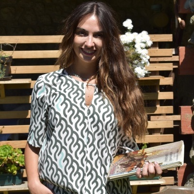 Mulher usando blusa padrão ondas preto e branco segurando revista em ambiente exterior com plantas e pedra.