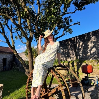 Mulher de vestido branco e chapéu de palha sentada em estrutura metálica circular num jardim
