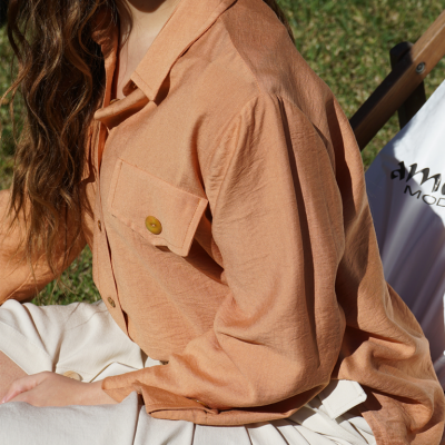 Blusa terracota com bolso e calças claras sentada em ambiente exterior com cadeira