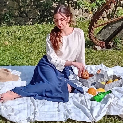 Mulher sentada num cobertor branco na relva com roupa branca e azul, comida e bebida à volta.