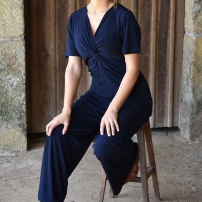 Mulher usando macacão azul escuro sentada em banco de madeira em ambiente rústico