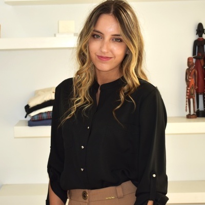 Mulher com blusa preta e calças bege em ambiente interno com prateleiras e esculturas