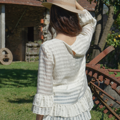 Mulher de costas com blusa branca de listras transparentes e chapéu bege em jardim