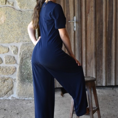 Mulher com macacão azul escuro de manga curta e pernas largas junto a porta rústica de madeira