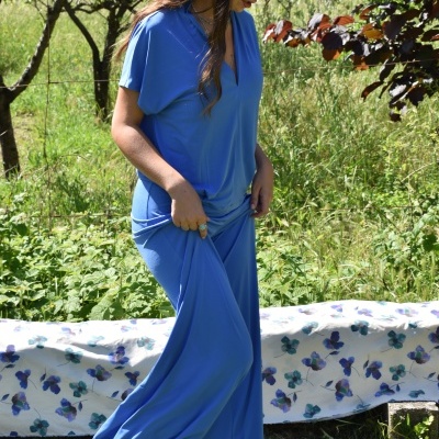 Mulher com conjunto de pijama azul claro de tecido leve num jardim, ao lado de um banco com cobertor floral azul.