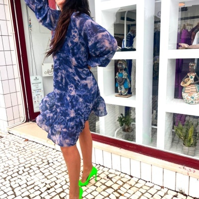 Mulher com vestido azul tie-dye, sapatos verdes de salto alto e óculos grandes, em calçada mosaico branco e preto em frente a loja