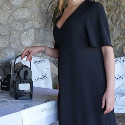 Mulher com vestido preto longo a tocar num objeto preto num ambiente com parede de pedra e mesa branca