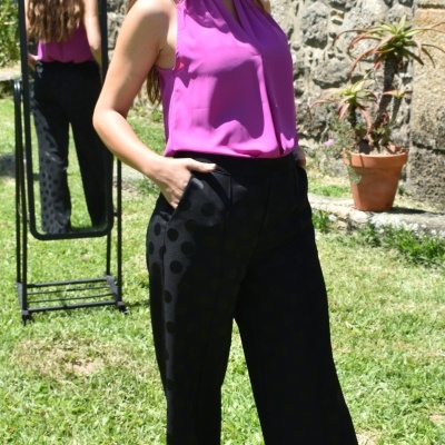 Mulher com blusa rosa e calças pretas com padrão de bolinhas em jardim com espelho e vaso