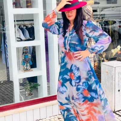 Mulher vestida com vestido longo colorido e chapéu fúcsia em frente a montra com manequins