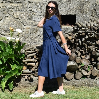 Mulher de vestido azul midi e sapatilhas brancas em jardim com parede de pedra e lenha