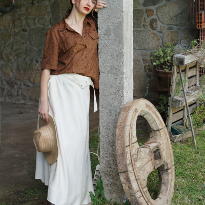 Mulher com blusa castanha, saia branca e chapéu de palha encostada em pilar com roda de madeira ao lado