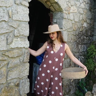 Mulher com macacão castanho às bolas brancas, chapéu de palha e cesto de vime junto a parede de pedra num jardim