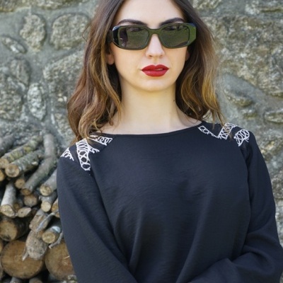 Mulher com óculos de sol negros e blusa preta com bordados brancos em frente a parede de pedra