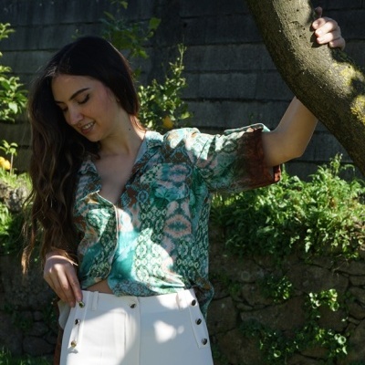 Mulher com camisa verde e calças brancas encostada a uma árvore num jardim