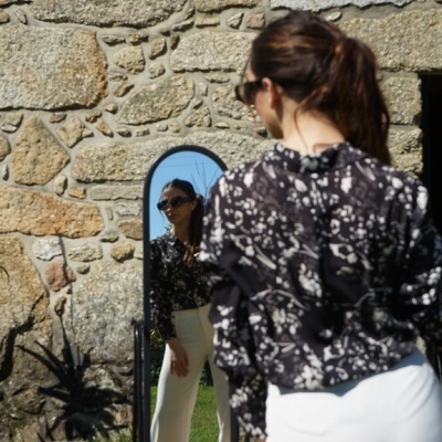 Mulher de costas com blusa preta estampada e calças brancas olhando para espelho em jardim com parede de pedra.