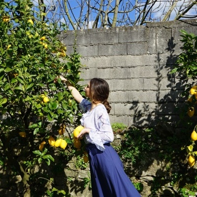 Mulher a apanhar limões num jardim com blusa branca e saia azul