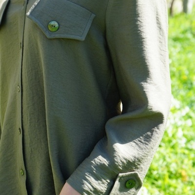 Camisa verde-oliva com botões, bolsos abotoados e fundo com vegetação