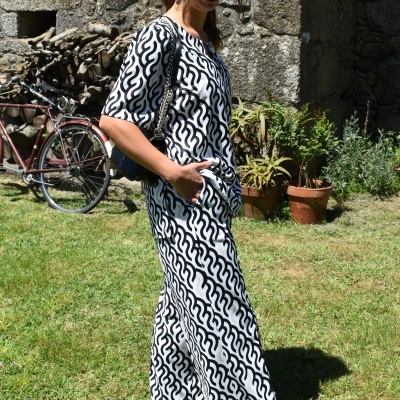 Mulher com conjunto branco e preto com padrão ondulado, óculos escuros e ténis castanhos em relva junto a parede de pedra