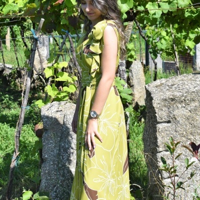 Mulher usando vestido longo verde com padrão floral, ao ar livre entre vinhas e pedras