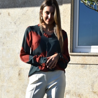 Mulher com blusa verde escuro e terracota e calças brancas em parede de pedra.