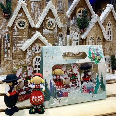 Bonecos de Natal em madeira com roupa tradicional em caixa decorativa junto a casas de madeira com neve.