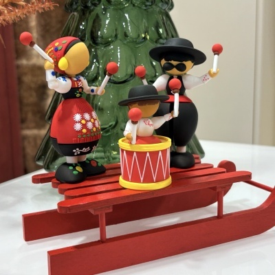 Três figuras de madeira vestidas a rigor em trajes tradicionais com instrumentos num trenó vermelho.