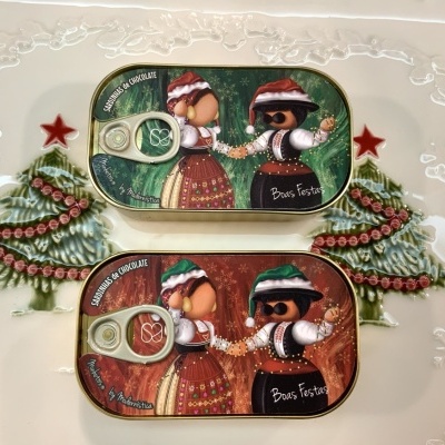 Duas latas de metal decoradas com ilustrações natalícias e texto, sobre uma superfície com decoração de árvores de Natal.