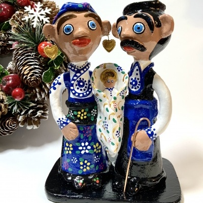 Escultura de casal e criança em cerâmica com trajes tradicionais coloridos, sobre base preta, com enfeite natalício ao fundo.