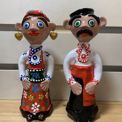 Duas figuras de cerâmica colorida vestidas com trajes tradicionais, mulher e homem, em cima de mesa de madeira clara.