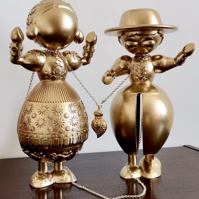 Duas figuras metálicas douradas decorativas, uma feminina e outra masculina, ligadas por corrente com coração