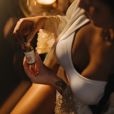 Mulher segurando frasco pequeno com conta-gotas, roupão branco com renda, ambiente interior com velas acesas