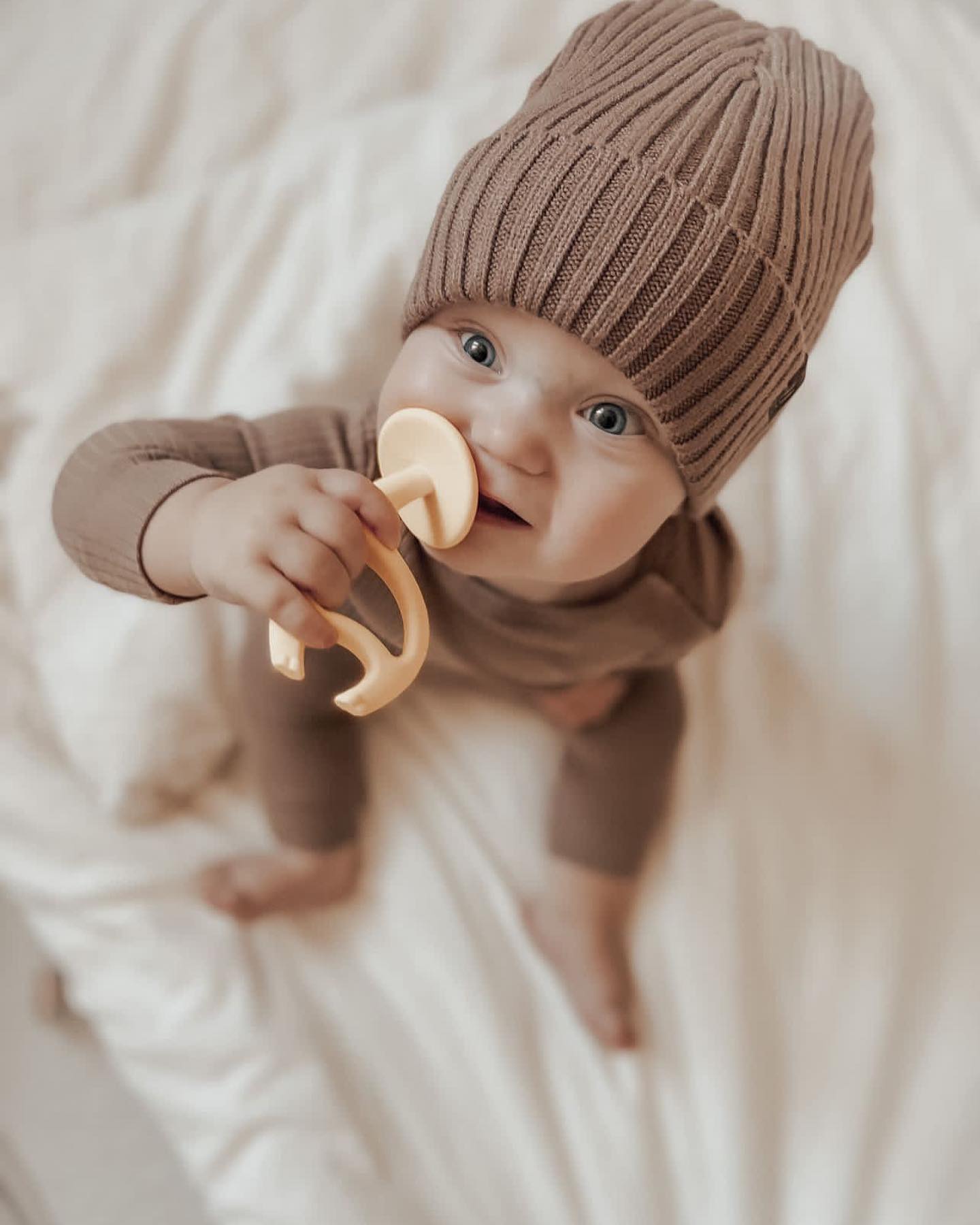 Bebé com gorro castanho e anel de dentição amarelo claro em fundo branco