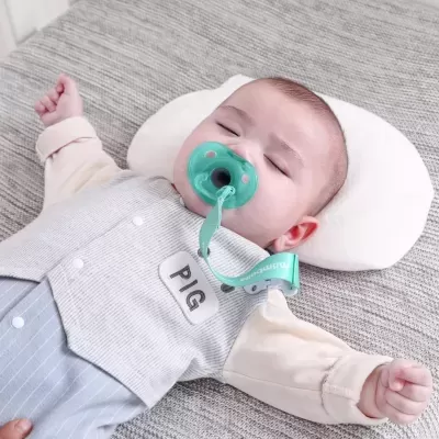 Bebé a dormir com chupeta verde e roupa cinza e azul deitado numa almofada branca