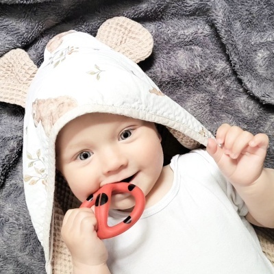 Bebé com toalha branca com orelhas castanhas a morder mordedor vermelho com pontos pretos, sobre superfície cinzenta macia.