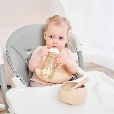 Bebé numa cadeira alta a beber numa garrafa e com uma tigela e colher à sua frente