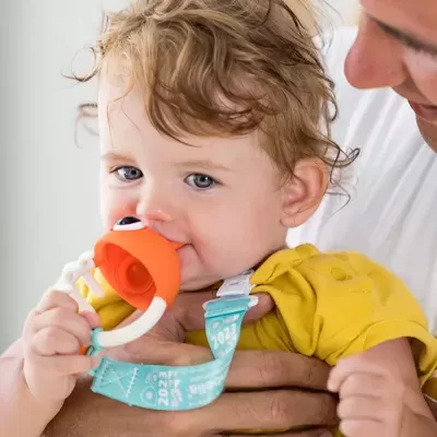 Bebé com brinquedo laranja de silicone e suporte azul claro, vestido com camisa amarela, segurado por adulto