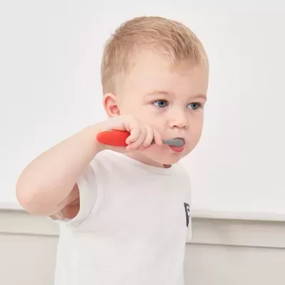 Criança loira escovando os dentes com escova infantil vermelha e cinza, vestindo t-shirt branca