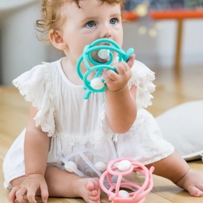 Bebé sentada no chão segurando brinquedo de silicone azul, com outro rosa ao lado