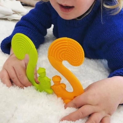 Criança com suéter azul segurando brinquedos de silicone verde e laranja em superfície branca