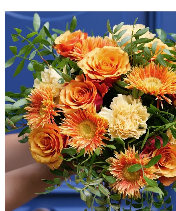 Bouquet de flores com rosas laranja, cravos creme e flores laranja e amarelas, folhagem verde, fundo azul