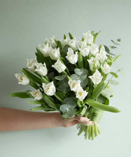 Bouquet de flores brancas com folhas verdes e ramos de eucalipto, mão segurando, fundo cinza claro