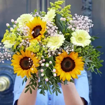 Ramo de flores com girassóis, flores brancas e botões rosa em fundo desfocado.