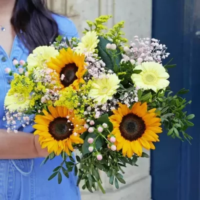 Ramo colorido de girassóis e flores diversas com folhagem verde, segurado por pessoa em vestido azul claro