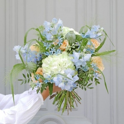 Buquê de flores azuis, brancas e pêssego com folhas verdes, segurado por uma mão com manga branca